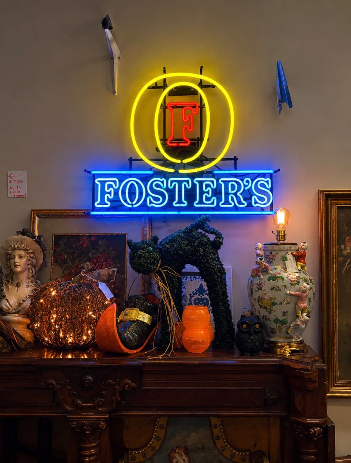 Foster’s neon sign above a carved antique mantel with a porcelain lamp and vintage Halloween decor